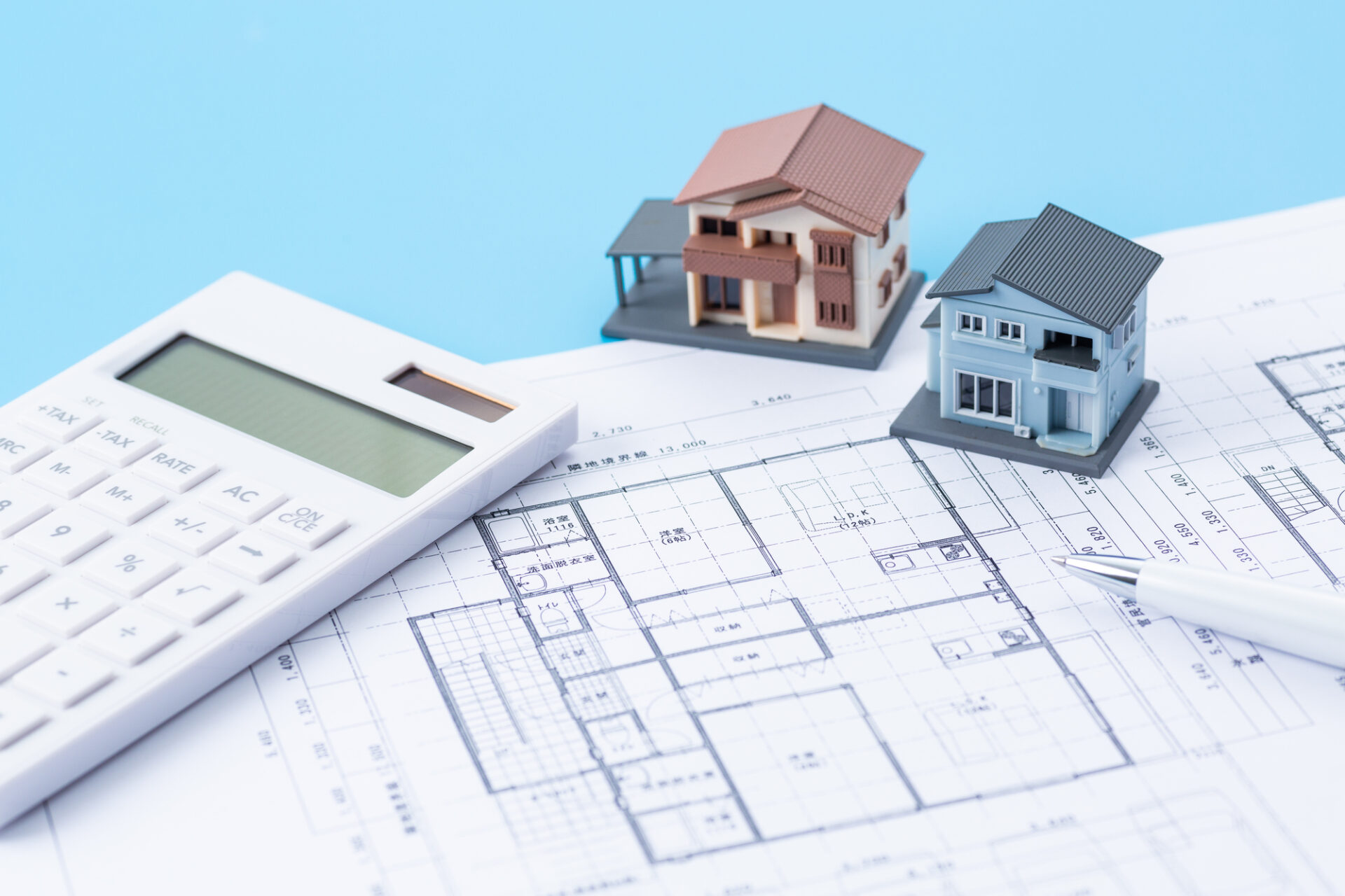 間取り図の上に置かれた電卓と住宅模型2棟、住宅建築費用の計算や資金計画のイメージ
